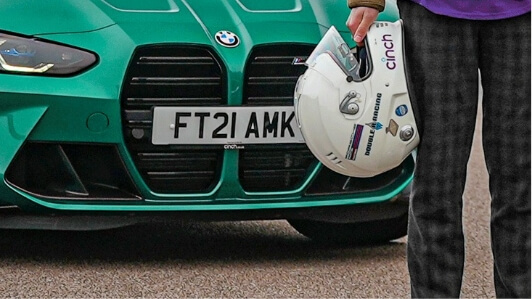 A person standing in front of a BMW car holding a racing helmet with a cinch sponsor